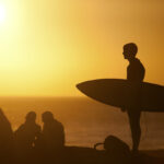 Surfer carrying surfboard on beach at sunset, Taghazout via Depositphotos