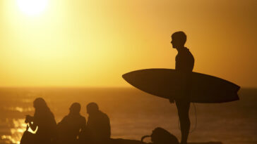 Surfer carrying surfboard on beach at sunset, Taghazout via Depositphotos