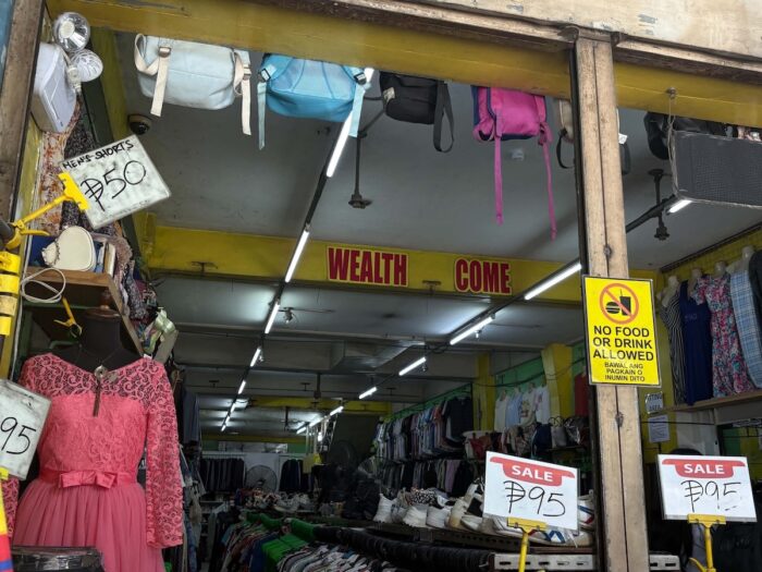 A snapshot of daily commerce: Bargain signs, hanging backpacks, rack of clothes, and friendly reminders at a local thrift shop storefront.